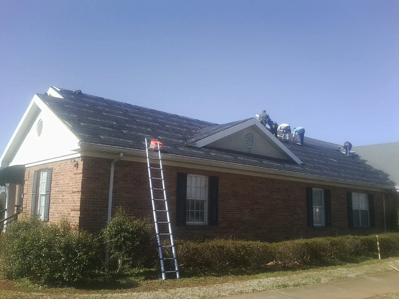 Crew tearing shingles off a brick ranch roof with a ladder set — Emergency Roof Repair work by our crew in Nedrow