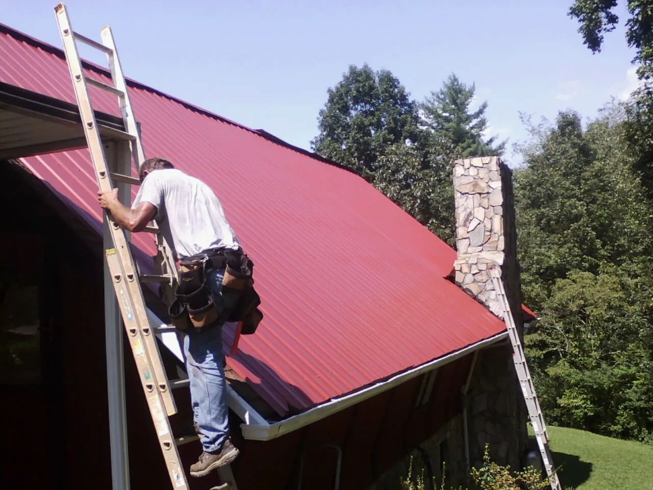 Roofer on a ladder beside a red metal roof and stone chimney — Soffit & Fascia Repair by Roofing Brothers in Nedrow